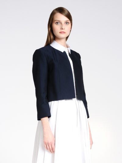 Une mannequin aux cheveux longs et bruns porte une robe blanche à col et une veste courte bleu marine. La veste est dotée de manches longues et d'un devant ouvert. Elle se tient debout sur un fond blanc uni, regardant directement l'objectif, dégageant le charme de la mode parisienne dans cette séance photo chic réalisée par un photographe de renom.