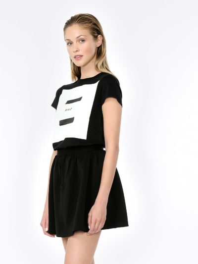 Une mannequin aux cheveux blonds, ressemblant à une icône de la mode parisienne, se tient debout sur un fond clair et uni. Elle porte un t-shirt graphique noir et blanc associé à une jupe noire, la tête légèrement inclinée sur le côté et les bras pendants le long du corps. La photo respire un chic décontracté typique de la photographie de rue à Paris.