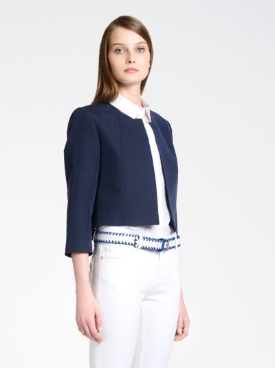 Une mannequin se tient debout sur un fond blanc uni, vêtue d'une veste courte bleu marine sur une chemise blanche boutonnée et d'un pantalon blanc avec une ceinture tissée bleue et blanche. Ses cheveux raides sont séparés sur un côté et tombent sur son épaule tandis qu'elle regarde légèrement hors champ, incarnant la mode parisienne à la Thomas Duclert.