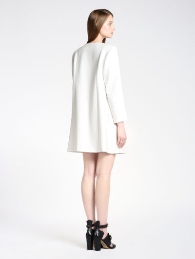 Une personne aux cheveux longs et raides se tient debout et regarde légèrement vers la droite. Elle porte une robe blanche à manches longues qui lui arrive aux genoux. Elle porte des chaussures noires à talons hauts avec des brides aux chevilles. L'arrière-plan est blanc uni, une photo classique qui s'intégrerait parfaitement dans n'importe quel portfolio de photographe de mode de Thomas Duclert.