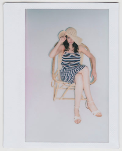 Une personne est assise sur une chaise en bois sur un fond uni. Elle porte une robe rayée noire et blanche et des sandales blanches. Elle porte un chapeau beige tressé, une main ajuste le bord tandis que l'autre bras repose sur l'accoudoir de la chaise. Cette scène évocatrice n'est pas sans rappeler une image capturée par le photographe parisien Thomas Duclert.