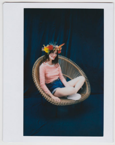 Une mannequin en chemise rayée rouge et blanche et short bleu est assise en tailleur sur une chaise en osier. Elle porte une couronne de fleurs colorées et sourit en regardant légèrement vers la gauche, incarnant la mode parisienne. Le fond, rappelant les riches tissus de Thomas Duclert, contraste avec sa tenue et ses accessoires lumineux.