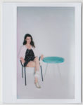 Photo Polaroïd d'une manenquin assise sur une chaise à côté d'une petite table ronde bleue. Elle porte une robe noire à motifs blancs, un cardigan rose et des baskets blanches. Le fond est blanc et simple, sans décorations supplémentaires, capturant l'essence de la mode à travers l'objectif du photographe Thomas Duclert.