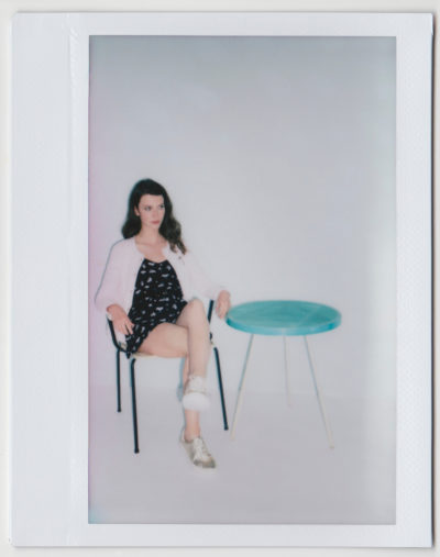 Photo Polaroïd d'une manenquin assise sur une chaise à côté d'une petite table ronde bleue. Elle porte une robe noire à motifs blancs, un cardigan rose et des baskets blanches. Le fond est blanc et simple, sans décorations supplémentaires, capturant l'essence de la mode à travers l'objectif du photographe Thomas Duclert.
