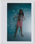 Sur une photo Polaroïd du photographe Thomas Duclert, une mannequin se tient debout, confiante, vêtue d'une robe rose sur un haut blanc à manches longues. Elle pose les mains sur les hanches sur un fond bleu aux motifs lumineux tachetés, incarnant un style décontracté parfaitement associé à des baskets.
