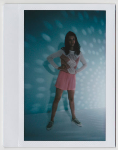 Sur une photo Polaroïd du photographe Thomas Duclert, une mannequin se tient debout, confiante, vêtue d'une robe rose sur un haut blanc à manches longues. Elle pose les mains sur les hanches sur un fond bleu aux motifs lumineux tachetés, incarnant un style décontracté parfaitement associé à des baskets.