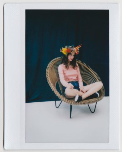 Une personne aux cheveux longs, coiffée d'une couronne de fleurs, d'un haut à manches longues à rayures roses et blanches et d'un short en jean, est assise en tailleur sur une chaise en osier devant un fond bleu foncé. Prise par le photographe Thomas Duclert, l'image présente une bordure blanche de style Polaroïd.