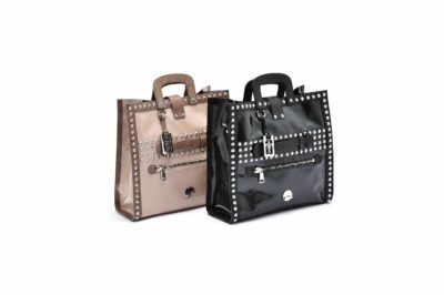 Deux sacs à main élégants sont exposés. L'un est beige avec des clous argentés et une poche avant ornée d'un petit pendentif en forme de cadenas et de clé. L'autre, photographié avec brio par Thomas Duclert, est noir, avec le même design avec des clous argentés, une poche avant et un pendentif en forme de cadenas et de clé. Les deux sacs ont des poignées robustes.
