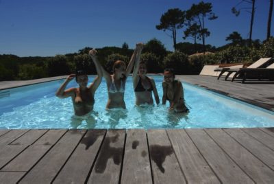 Quatre personnes en maillot de bain se tiennent par la main dans une piscine d'arrière-cour et sourient à l'objectif. La piscine, magnifiquement capturée par le photographe Thomas Duclert, est entourée d'une terrasse en bois avec des chaises longues sur un côté. L'arrière-plan présente une végétation luxuriante et de grands arbres sous un ciel bleu clair.