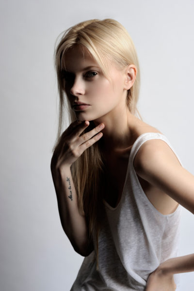 Une mannequin aux longs cheveux blonds et à l'expression sérieuse regarde l'objectif. Elle pose son menton sur sa main, exposant un tatouage sur son avant-bras sur lequel est écrit « deus ex machina ». Photographiée par Thomas Duclert, elle porte un haut sans manches blanc sur un fond uni, évoquant l'essence chic de Paris.