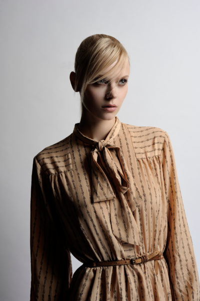 Une personne aux cheveux blonds coiffés de façon élégante et balayée sur le côté porte une robe marron clair à manches longues avec un détail de col noué et une fine ceinture à la taille. Le photographe Thomas Duclert capture cette mode sur un fond uni gris clair.