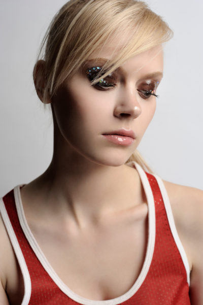 Gros plan d'une personne aux cheveux blonds clairs tirés en arrière, portant un maquillage léger avec un fard à paupières pailleté. Elle est vêtue d'un débardeur rouge à bordure blanche, le regard baissé et légèrement sur le côté, sur un fond gris clair uni, capturant l'essence de la mode parisienne.