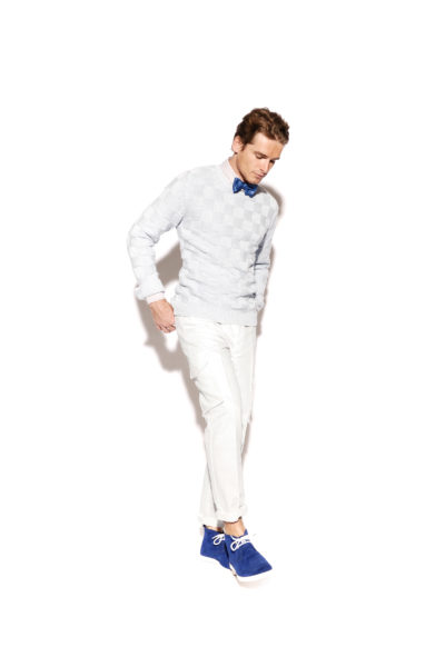 Une mannequin vêtue d'un pull à carreaux clair, d'un pantalon blanc et de chaussures bleu vif à lacets blancs pose sur un fond blanc uni. Avec un nœud papillon bleu et une main dans la poche, elle incarne l'élégance stylée capturée par le photographe de mode Thomas Duclert.