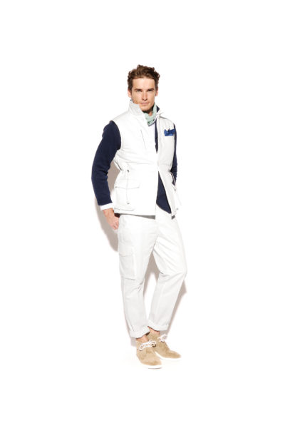 Thomas Duclert pose sur un fond blanc uni, incarnant la mode parisienne avec son gilet blanc sans manches sur une chemise à manches longues foncée, un pantalon blanc et des chaussures beiges. Il complète son look chic avec un foulard léger autour du cou et un mouchoir bleu dans la poche de son gilet.