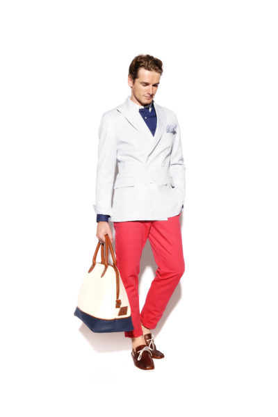 Un homme se tient debout sur un fond blanc, vêtu d'un blazer gris clair sur une chemise bleu marine, d'un pantalon rouge vif et de chaussures marron. Il tient dans sa main gauche un sac fourre-tout crème et bleu marine avec des poignées en cuir marron. Stylé comme s'il sortait des rues de Paris, son regard est dirigé vers le bas.