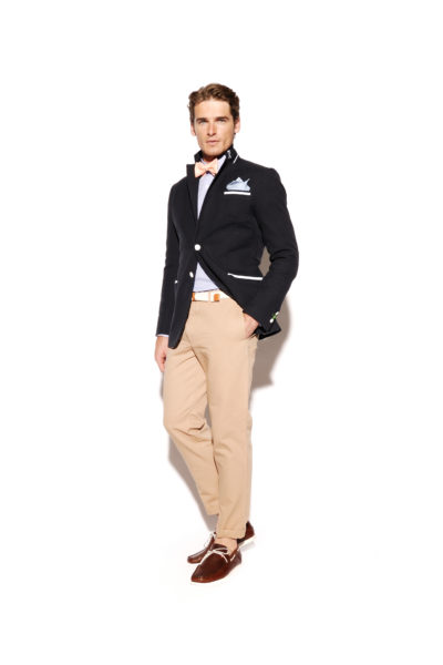 Un homme se tient debout, confiant, sur un fond blanc. Il porte un blazer bleu marine, une chemise bleu clair, un pantalon beige et des chaussures marron. Il accessoirise son look avec un nœud papillon rose, une ceinture blanche et une pochette. Dans la lignée de la mode parisienne, les mains de Thomas Duclert reposent nonchalamment dans ses poches.