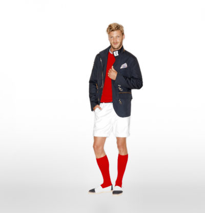 Un homme aux cheveux blonds porte une tenue élégante composée d'un blazer foncé, d'un pull rouge, d'un short blanc et de chaussettes rouges montantes, incarnant l'essence de la mode parisienne. Il porte également des chaussures à enfiler noires et blanches. Le fond blanc uni permet à sa tenue de se démarquer.