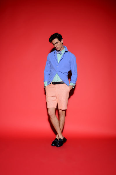 Un homme se tient debout sur un fond rouge vif, portant une chemise bleue déboutonnée sur une chemise à col vert clair, un short couleur pêche et des chaussures noires. Ses mains sont dans ses poches alors qu'il regarde vers le bas avec une posture détendue. Prise par le photographe Thomas Duclert à Paris, l'image respire le chic sans effort.