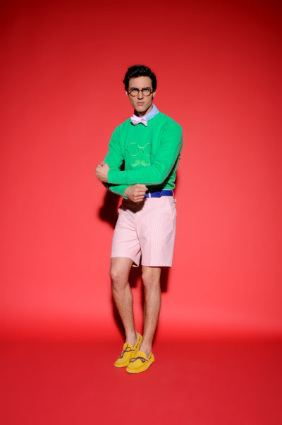 Un homme se tient debout sur un fond rouge vif, portant un sweat-shirt vert avec une illustration de lunettes, un nœud papillon rose, une montre blanche, un short rose pâle, une ceinture bleue et des chaussures jaunes. Il prend la pose avec son bras droit croisé sur sa poitrine, regardant directement l'objectif sur cette photographie de mode parisienne.