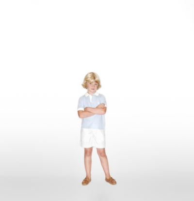 Un jeune enfant aux cheveux blonds courts se tient debout sur un fond blanc. L'enfant, photographié par Thomas Duclert à Paris, porte une chemise bleu clair, un short blanc et des chaussures marron, les bras croisés et une expression sérieuse.
