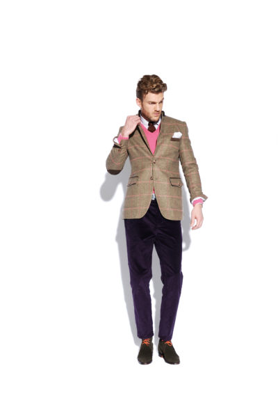 Un homme aux cheveux coiffés se tient debout sur un fond blanc uni, vêtu d'un blazer en tweed, d'un pull rose, d'une chemise blanche et d'un nœud papillon. Il porte un pantalon sombre et des chaussures à lacets, ajustant d'une main son blazer. Captures par le photographe Thomas Duclert, des ombres sont projetées sur l'arrière-plan.
