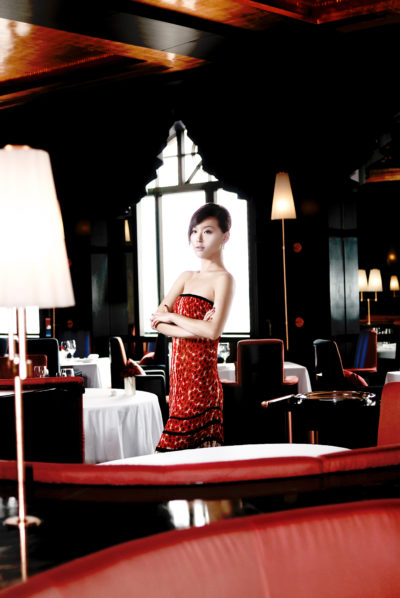 Une mannequin vêtue d'une robe bustier rouge se tient debout, les bras croisés, dans un restaurant élégant et faiblement éclairé. La salle à manger, qui rappelle le chic parisien, présente une décoration moderne avec des murs noirs et de grandes fenêtres. Les tables sont dressées avec des nappes blanches et des chaises élégantes, rouges et noires.