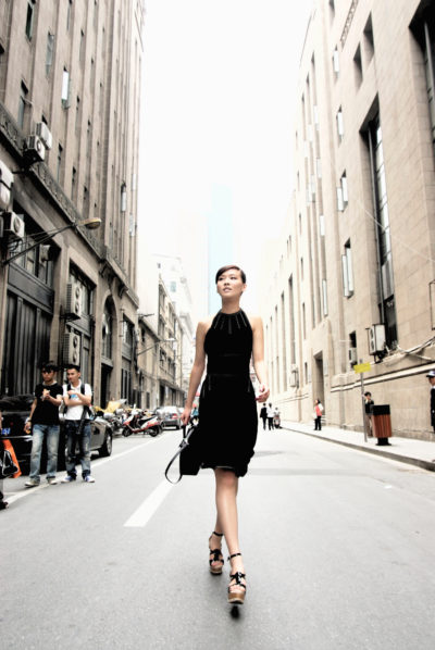 Une mannequin en robe noire et talons hauts marche avec assurance dans une rue parisienne bordée de hauts immeubles. Elle porte un petit sac noir et regarde droit devant elle, comme si elle sortait d'une séance photo de mode, tandis que des piétons et des voitures garées sont visibles en arrière-plan par une journée légèrement nuageuse.