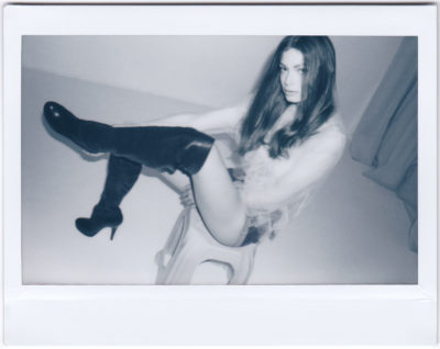 Une photo en niveaux de gris prise par un photographe parisien présente une mannequin aux cheveux longs, portant un haut transparent et des bottes à talons hauts. Elle prend la pose sur un tabouret avec une jambe surélevée et pliée au niveau du genou. L'arrière-plan minimaliste met en valeur le style chic.