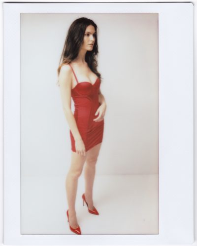 Une mannequin se tient debout sur un fond uni, vêtue d'une robe rouge ajustée sans manches et de talons hauts rouges. Avec son air de mode parisienne, elle a de longs cheveux noirs et regarde sur le côté avec une expression neutre, comme si elle avait été capturée par un photographe dans un instant fugace.