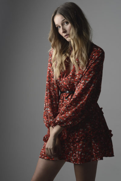 Une mannequin aux cheveux blonds longs et ondulés porte une robe rouge à motifs floraux. Elle pose les bras légèrement pliés et affiche une expression douce sur son visage, rappelant la mode parisienne. La photographie, prise par Thomas Duclert, présente un fond gris neutre.