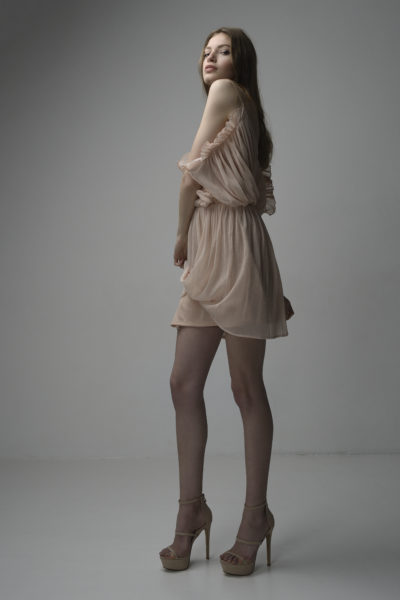 Une mannequin aux cheveux longs et bruns pose dans une robe beige clair à épaules dénudées. Elle se tient debout sur des sandales à talons hauts et regarde légèrement par-dessus son épaule avec une expression neutre. Capturée par le photographe Thomas Duclert, le fond gris uni donne à l'image une touche minimaliste.