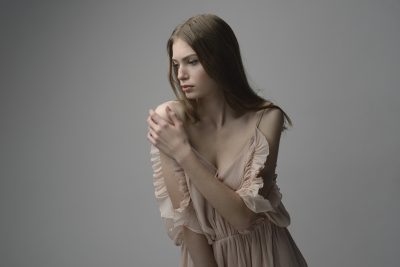 Une mannequin aux cheveux longs et raides, bruns, vêtue d'une robe rose pâle à volants, pose sur un fond gris neutre. Elle touche doucement son épaule gauche avec sa main droite et regarde vers la droite avec une expression pensive, incarnant l'élégance de la mode parisienne sous l'objectif d'un photographe talentueux.