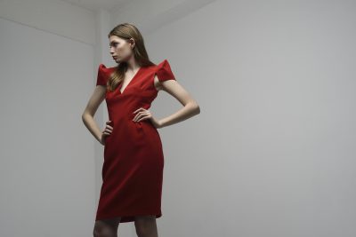 Une mannequin en robe rouge se tient debout, les mains sur les hanches, sur un fond gris clair uni. Elle a les cheveux longs et raides et regarde sur le côté. La robe présente des manches courtes et structurées et un décolleté en V. Prise par le photographe Thomas Duclert à Paris, la scène semble se dérouler en intérieur.