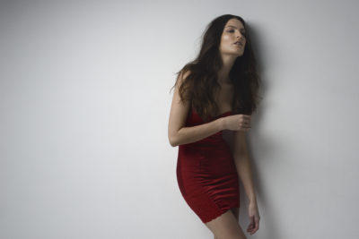 Une mannequin aux longs cheveux noirs porte une robe moulante rouge sur un fond blanc uni. Photographiée par Thomas Duclert, elle se penche légèrement sur le côté, le regard levé avec une expression détendue. Son bras est plié au niveau du coude et ses doigts touchent sa taille, incarnant l'élégance parisienne.