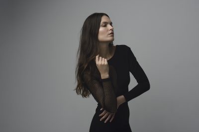 Une mannequin aux cheveux longs et foncés porte une robe noire ajustée aux manches en filet et pose devant un fond gris uni. Photographiée par Thomas Duclert, elle se tient debout, une main posée sur son ventre et l'autre près de son visage, les yeux fermés dans une expression sereine.