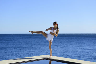 Une mannequin vêtue d'une tenue de sport blanche exécute un coup de pied haut sur une plate-forme au bord d'une piscine réfléchissante avec un vaste océan en arrière-plan sous un ciel bleu clair, comme si elle était prête pour une séance photo de mode haute couture par un photographe parisien.