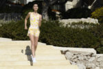 Une mannequin en tenue de sport, comprenant un débardeur à motifs jaune et blanc et un short assorti, monte un escalier en pierre à la parisienne. L'arrière-plan comprend de la verdure, des murs en pierre et une partie d'un bâtiment. Elle semble concentrée et active.