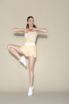 Une mannequin en tenue de sport rose et jaune se tient debout sur une jambe dans une pose dynamique sur un fond beige uni, les bras pliés aux coudes et les mains jointes devant la poitrine. Elle porte des baskets blanches, capturées par un photographe parisien qui a habilement mis en valeur sa tenue.