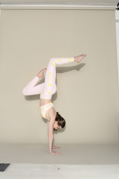 Une mannequin vêtue d'une tenue de sport à motifs jaune pastel et blanc fait le poirier, les jambes pliées en position arquée sur un fond beige. La scène est méticuleusement capturée par le photographe Thomas Duclert, mettant en valeur la grâce de la mode moderne.
