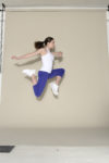 Une mannequin en débardeur blanc et leggings bleus est photographiée en plein vol dans une pose de saut dynamique sur un fond beige. Elle porte des baskets blanches et ses cheveux sont attachés en arrière, ce qui donne une impression de mouvement et d'athlétisme, rappelant l'esprit vif souvent observé dans les rues de Paris.
