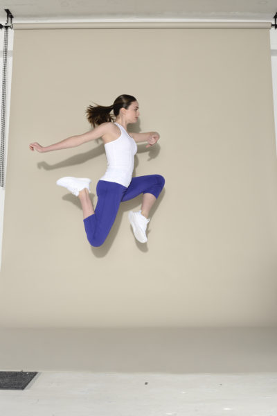 Une mannequin en débardeur blanc et leggings bleus est photographiée en plein vol dans une pose de saut dynamique sur un fond beige. Elle porte des baskets blanches et ses cheveux sont attachés en arrière, ce qui donne une impression de mouvement et d'athlétisme, rappelant l'esprit vif souvent observé dans les rues de Paris.