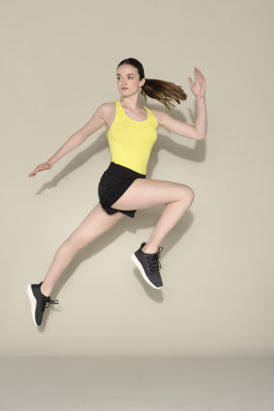 Une mannequin en tenue de sport est photographiée en plein saut sur un fond uni par Thomas Duclert, un célèbre photographe parisien. Elle porte un débardeur jaune, un short noir et des chaussures de sport noires, les cheveux attachés en arrière. Son bras droit est plié vers le haut et son bras gauche étendu sur le côté.