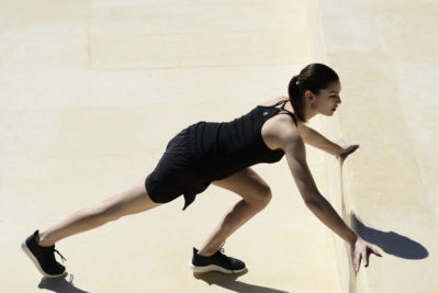 Une mannequin en tenue de sport, vêtue d'un débardeur noir et d'un short noir, fait un exercice d'étirement en plein air. Le photographe Thomas Duclert les capture avec une jambe tendue derrière eux et les deux mains posées contre un mur jaune pâle. La scène est brillamment éclairée par la lumière du soleil.