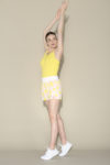 Une mannequin vêtue d'un débardeur jaune, d'un short à motifs jaune et blanc et de baskets blanches effectue un exercice d'étirement avec les bras levés au-dessus de la tête. Sur un fond beige uni, elle prend une pose rappelant celle d'un mannequin parisien photographié par un photographe.