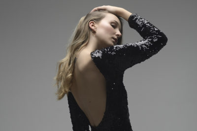 Une mannequin aux cheveux longs et ondulés pose avec sa main droite sur la tête. Elle porte une robe noire à paillettes et dos nu. Photographiée par Thomas Duclert à Paris, le fond gris uni met en valeur l'élégance et les détails de sa tenue.
