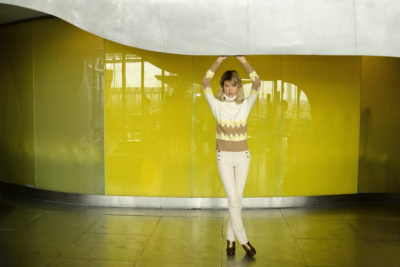 Une personne porte un pull jaune et blanc à motifs en zigzag, un pantalon clair et des chaussures marrons, pose devant un mur jaune et vert. Les bras levés au-dessus de la tête, l'arrière-plan intérieur met en valeur des éléments métalliques et vitrés, incarnant la mode parisienne inspirée par Thomas Duclert.