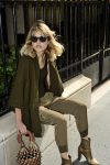 Une personne aux cheveux blonds portant des lunettes de soleil et un cardigan vert foncé est assise sur un rebord de pierre près d'une balustrade noire à Paris. Elle est vêtue d'un haut noir, d'un pantalon kaki et de bottes à talons marron clair, tenant un sac à main tressé avec des poignées en bois, capturant ce mode sans effort que les photographes adorent.