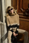 Une personne aux cheveux blonds portant des lunettes de soleil foncées et un pull à col roulé beige à motif en zigzag noir et blanc est assise contre un mur beige et une porte en bois. Rappelant le style parisien, ils sont également vêtus d'un pantalon beige et ont l'air détendu, capturant l'essence que l'on pourrait trouver dans une photographie de Thomas Duclert.