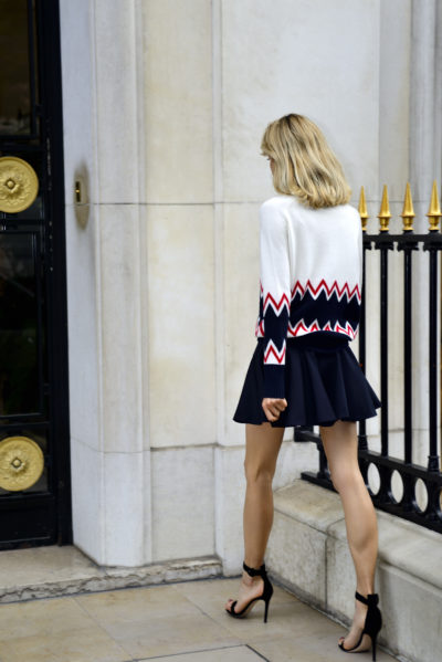 Une mannequin blonde, élégamment photographiée par Thomas Duclert, s'éloigne de l'objectif le long d'un bâtiment en pierre avec une clôture noire et dorée. Elle porte un pull blanc à manches longues avec des motifs en zigzag rouges et noirs, associé à une mini-jupe noire et à des sandales à talons hauts.