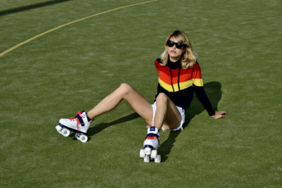 Une personne est assise sur l'herbe, elle porte des patins à roulettes, des lunettes de soleil et un haut rayé noir et rouge avec un short. Le soleil projette des ombres sur le sol et la personne semble détendue, avec une jambe pliée et l'autre tendue, ce qui donne un air décontracté souvent observé dans l'œuvre de Thomas Duclert.